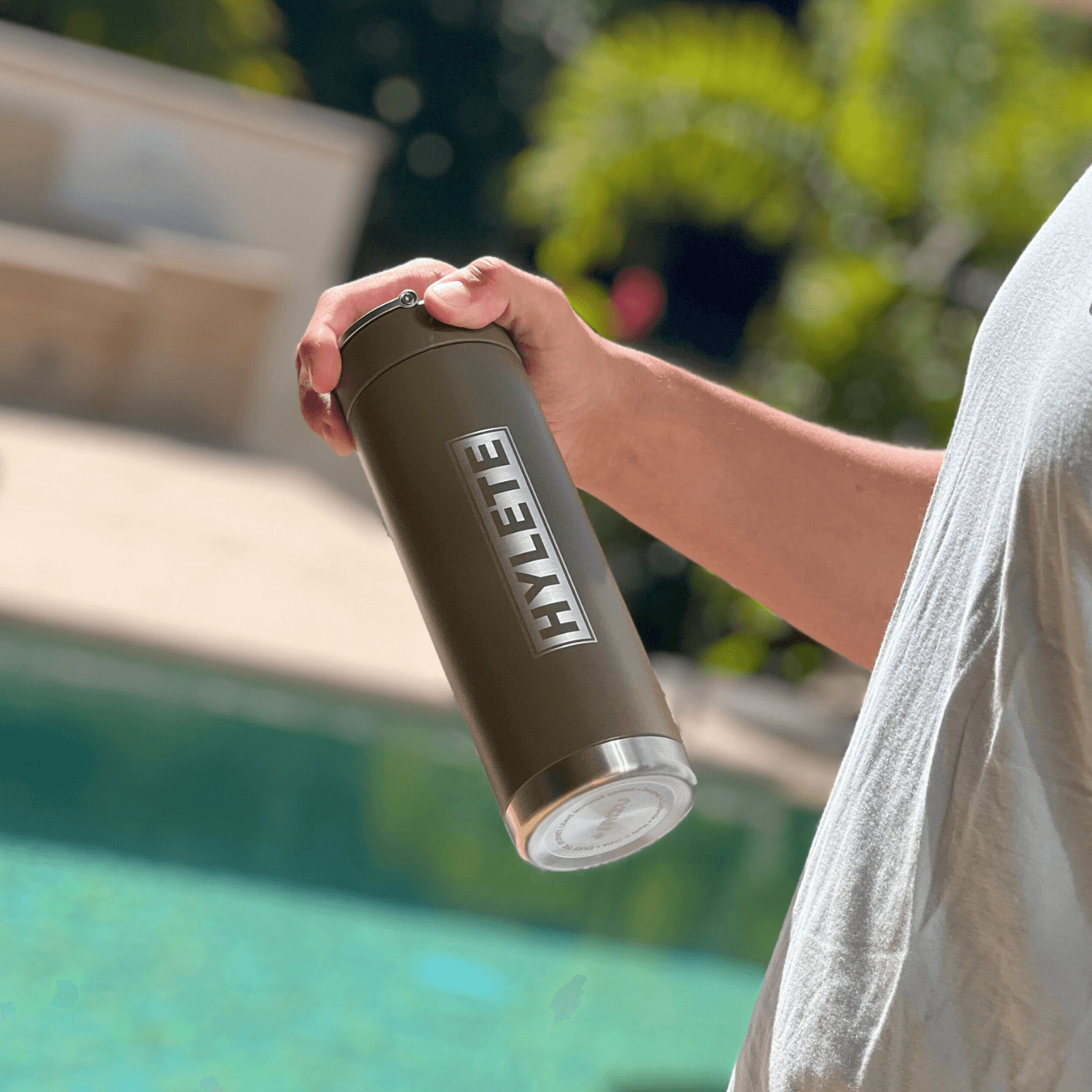 Person holding the HYLETE x Mizu Insulated Water Bottle in olive green by a pool, showcasing its sleek design and logo.